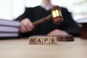 A judge holds a gavel behind wooden letter blocks spelling “RAPE” in a courtroom setting.